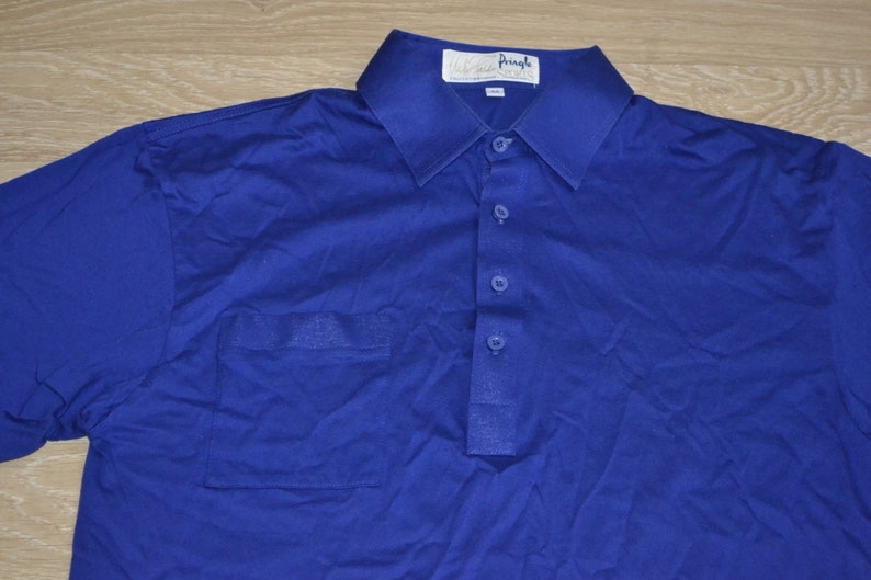 May include: A blue polo shirt with a single chest pocket and a button-up collar. The shirt has a white label with the text "Walter Pringle" on it.