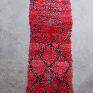 May include: A red Moroccan rug with a diamond pattern in green and black. The rug has a fringe on the bottom.