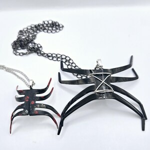 May include: Two black spider-shaped pendants on silver chains. The larger pendant has a silver wire design across the body. The smaller pendant has red and white paint splatters.