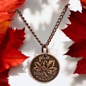 May include: A copper necklace with a 1955 Canadian 1 cent coin pendant featuring a maple leaf design. The coin is surrounded by red autumn leaves.