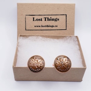 May include: A pair of cufflinks in a brown box with white tissue paper. The cufflinks are made from 1952 Canadian pennies, featuring a maple leaf design and the text "1 CENT 1952 CANADA".