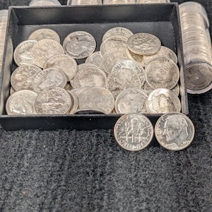 May include: A collection of silver dimes, some loose in a black tray and others in clear plastic tubes. The dimes feature the profile of a man and the words "United States of America" and "One Dime".