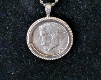 Op de afbeelding: Een zilverkleurige hanger met een 1964 Kennedy halve dollar munt in een gedraaide, touwachtige metalen rand. De munt toont het profiel van John F. Kennedy met de tekst IN GOD WE TRUST. Het hangt aan een zilveren ketting met kleine bolletjes.