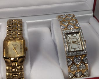 Op de afbeelding: Twee goudkleurige Elgin horloges in een wit satijn gevoerde doos. Eén horloge heeft een rechthoekige wijzerplaat en een gouden band. De andere heeft een rechthoekige wijzerplaat met een met diamant bezette band.