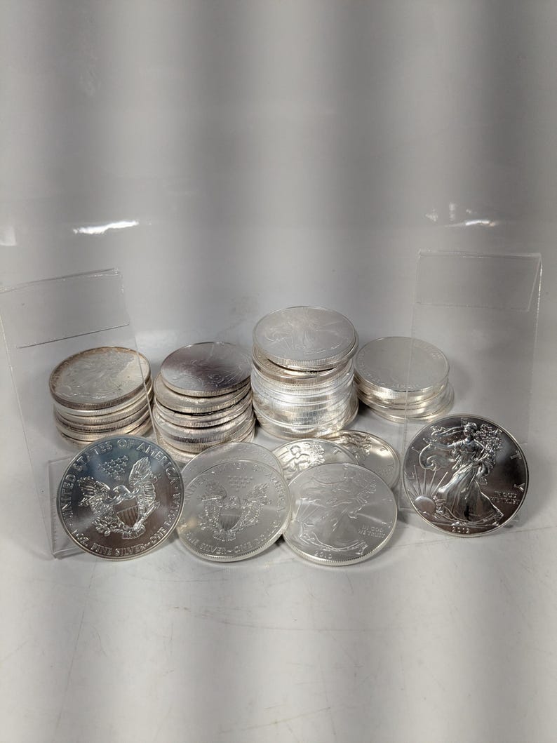 Op de afbeelding: Een verzameling zilveren American Eagle munten van &eacute;&eacute;n dollar, deels gestapeld en deels afzonderlijk weergegeven. De munten tonen het Walking Liberty-ontwerp aan de ene kant en een heraldische adelaar op de achterzijde, met zilveren oppervlakken.