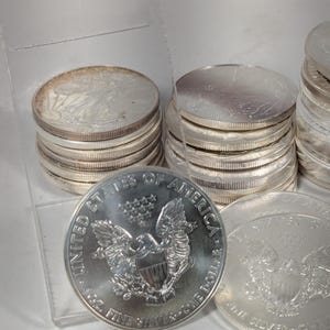 Op de afbeelding: Een verzameling zilveren American Eagle munten gestapeld in meerdere stapels. De voorste munt toont een adelaar met een schild, dertien sterren en de tekst United States of America, 1 oz. Fine Silver en One Dollar. De munten hebben een metallic afwerking.
