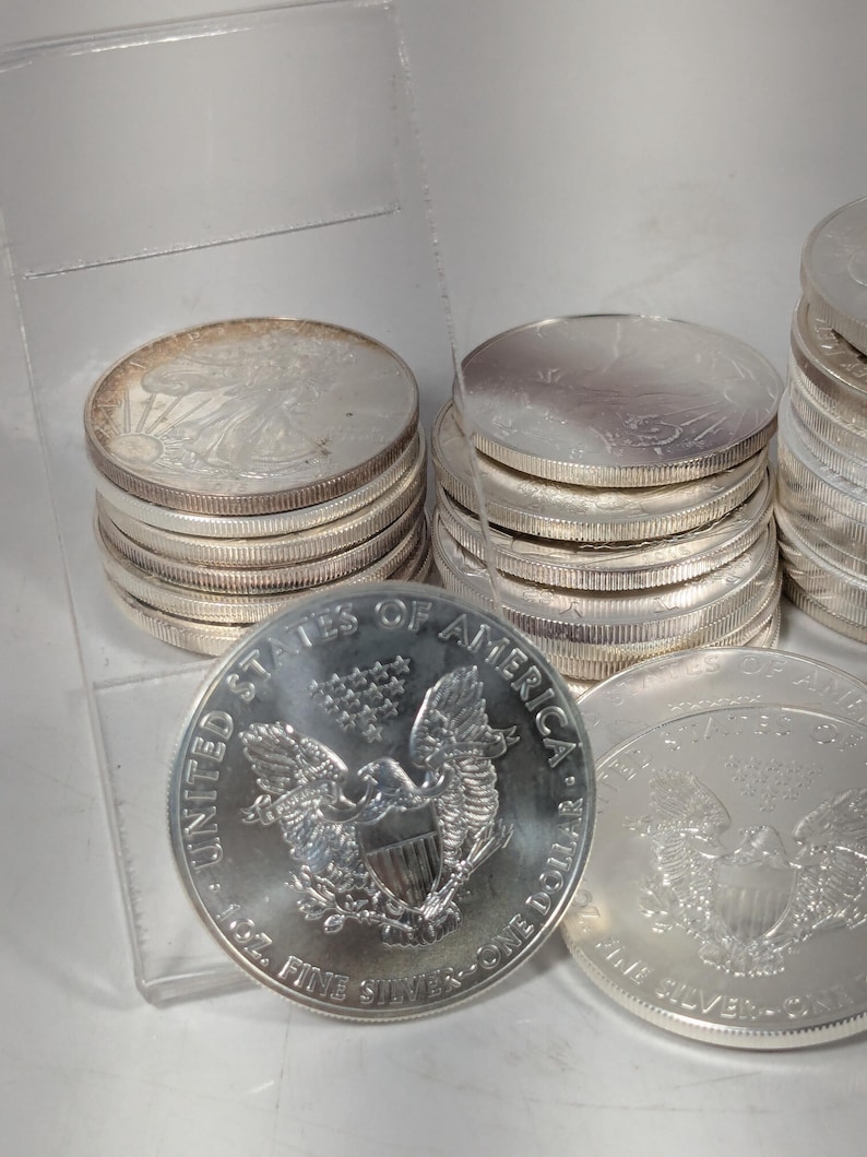 Op de afbeelding: Een verzameling zilveren American Eagle munten gestapeld in meerdere stapels. De voorste munt toont een adelaar met een schild, dertien sterren en de tekst United States of America, 1 oz. Fine Silver en One Dollar. De munten hebben een metallic afwerking.