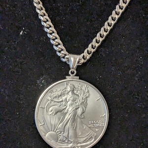 May include: Silver necklace with a large coin pendant. The coin shows a woman and the words "IN GOD WE TRUST" and the year 2022. The chain is a curb style, and the pendant is attached with a small bail.
