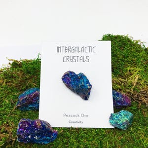 May include: A small piece of peacock ore, an iridescent blue and purple mineral, on a white card with the text "Intergalactic Crystals" and "Peacock Ore Creativity". The card is on a bed of green moss.