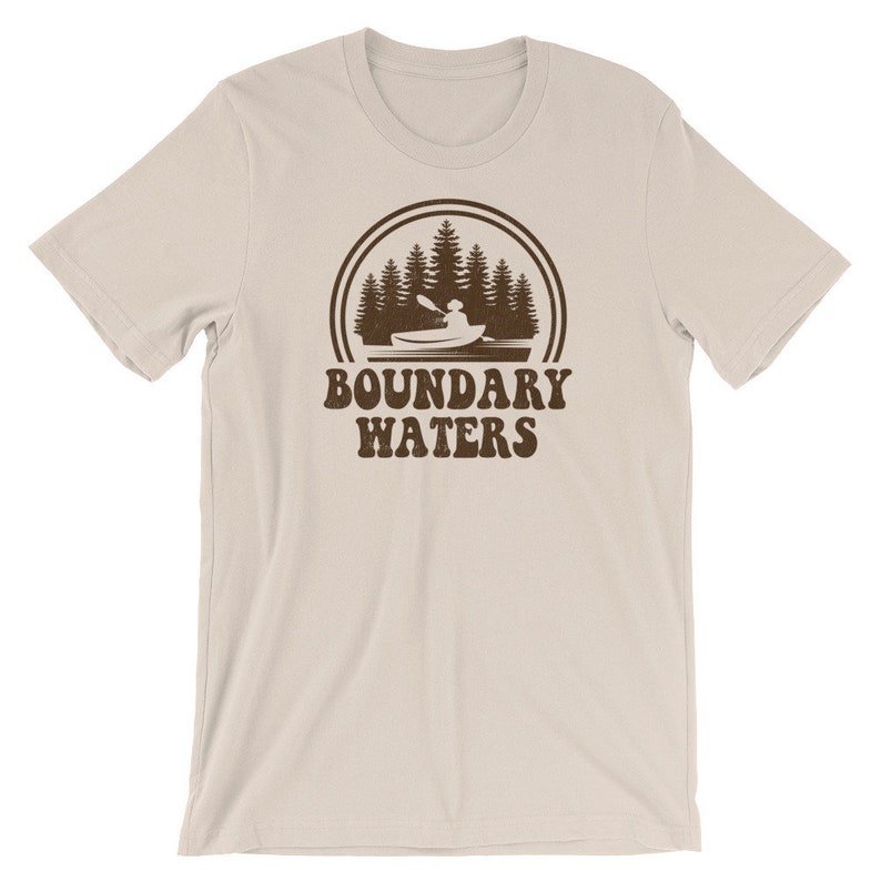 May include: A beige t-shirt with a brown graphic of a person kayaking in a lake surrounded by trees. The graphic is in a circle with the text "BOUNDARY WATERS" below it.