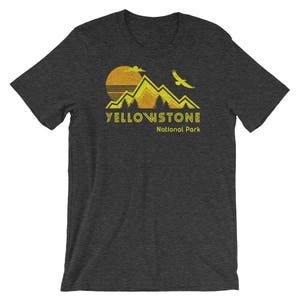 May include: A dark grey t-shirt with a yellow graphic of mountains, a sun, and a bird. The text "Yellowstone National Park" is printed in yellow below the graphic.