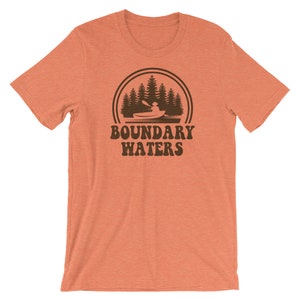 May include: Orange short-sleeved t-shirt with a brown graphic of a person kayaking in a lake with trees in the background. The graphic is surrounded by a circle with the text "Boundary Waters" underneath.