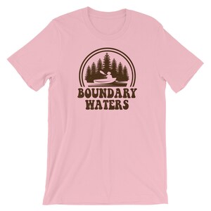 May include: Pink short-sleeved t-shirt with a brown graphic of a person kayaking in a lake with trees in the background. The graphic is surrounded by a circle with a double line border. The text "Boundary Waters" is printed below the graphic.