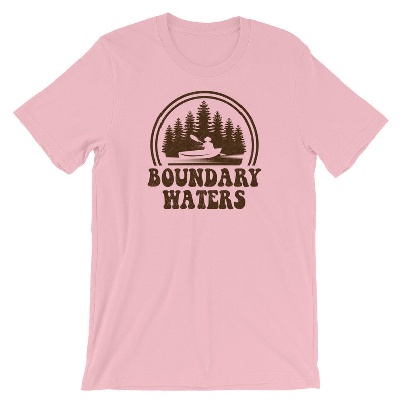May include: Pink short-sleeved t-shirt with a brown graphic of a person kayaking in a lake with trees in the background. The graphic is surrounded by a circle with a double line border. The text "Boundary Waters" is printed below the graphic.