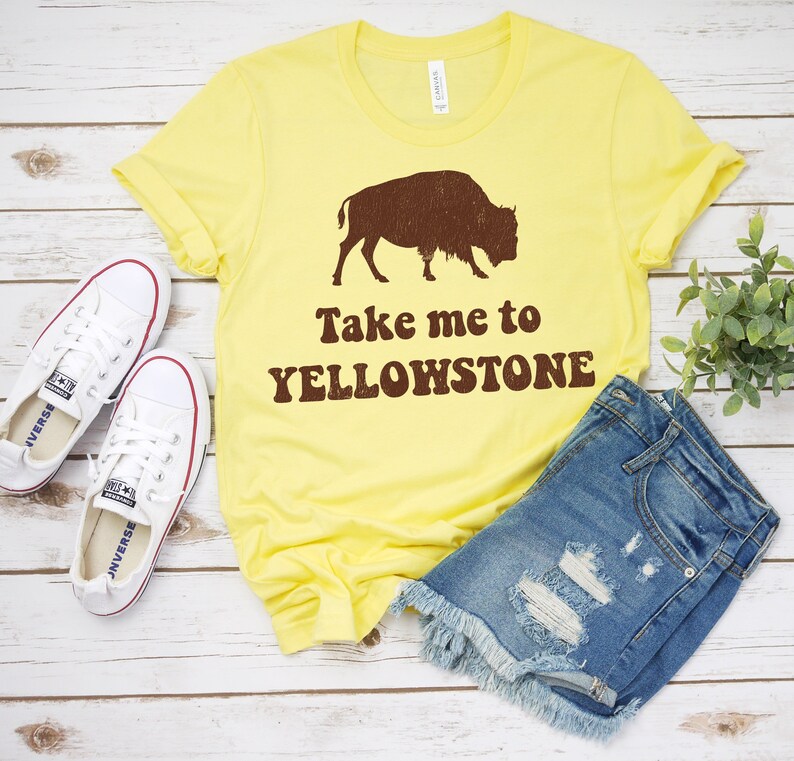 May include: Yellow t-shirt with a brown silhouette of a bison and the text "Take me to Yellowstone". The shirt is laid out on a wooden surface with a pair of white sneakers and denim shorts.