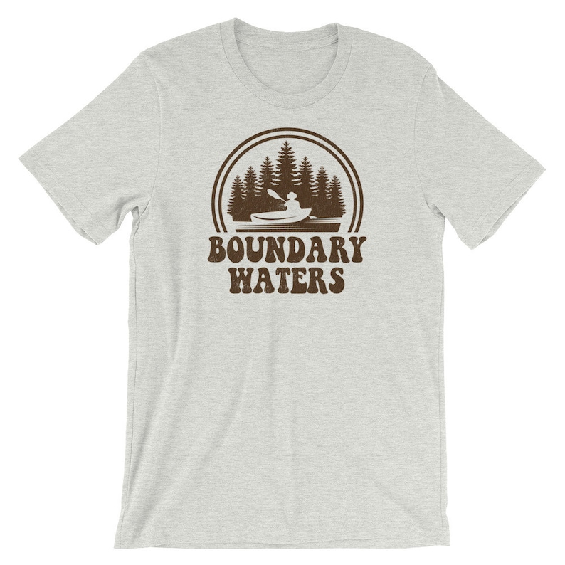 May include: A light gray t-shirt with a brown graphic of a person kayaking in a lake with trees in the background. The text "Boundary Waters" is below the graphic.