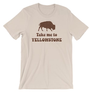 May include: A beige t-shirt with a brown silhouette of a bison and the text "Take me to Yellowstone" printed on the front.