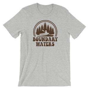 May include: A grey t-shirt with a brown graphic of a person kayaking in a forest with the text "Boundary Waters" underneath.