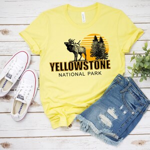 May include: Yellow t-shirt with a graphic of an elk, trees, and a sunset. The text "Yellowstone National Park" is printed in black on the shirt.