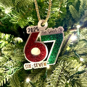 May include: A Christmas tree ornament with the text "2025 The Year Of 67 Six-Seven". The numbers 6 and 7 are red and green glitter. The ornament hangs from a gold cord against a backdrop of green Christmas tree needles.