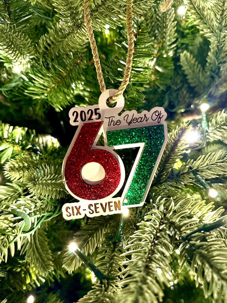 May include: A Christmas tree ornament with the text "2025 The Year Of 67 Six-Seven". The numbers 6 and 7 are red and green glitter. The ornament hangs from a gold cord against a backdrop of green Christmas tree needles.