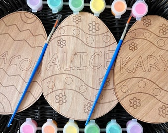 May include: Three wooden Easter egg shapes with paint-by-number designs. The eggs are labeled "ACO", "ALICE", and "KARY".  The eggs are surrounded by small containers of paint in various colors.