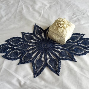 May include: A navy blue crocheted doily with a white ceramic trinket box with a floral design on top.