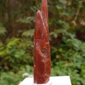 May include: A tall, pointed, red crystal with a rough, textured surface. The crystal is displayed on a white pedestal.