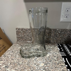 May include: A clear glass boot-shaped beer mug. The glass has a smooth, transparent surface and is designed to resemble a traditional boot. The mug is empty and sits on a speckled countertop.