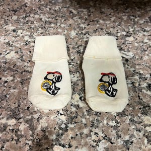 以下が含まれることがあります： テニスをしているスヌーピーの刺繍が施された白い足首丈の靴下2足セット。靴下の上部には赤と白のストライプのバンドが付いています。