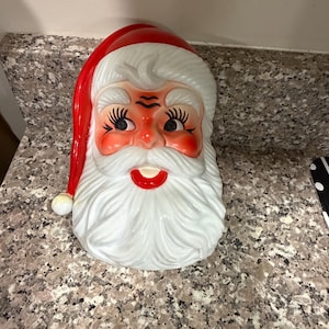 May include: A ceramic Santa Claus head with a red hat, rosy cheeks, and a white beard. The Santa head has black eyelashes and a red mouth. The Santa head is a festive decoration.