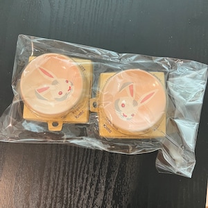 Könnte beinhalten: Zwei kleine, runde Keramikschalen mit rosa Hintergrund und einem weißen Hasendesign. Die Schalen sind in durchsichtigem Plastik verpackt.