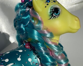 May include: A yellow My Little Pony with teal and pink hair. The pony has a blue and teal body with pink and white candy decorations and rhinestones.