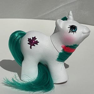 May include: A white unicorn figurine with a green mane and tail. The unicorn has a pink maple leaf on its side and a green and red design around its neck. The unicorn has blue eyes and a white horn.
