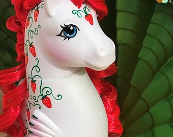 May include: A white toy pony with red hair and a strawberry design. The pony has blue eyes and pink cheeks. The strawberry design is on the pony's side and forehead. The pony is in a green shell-shaped container with colorful gems.