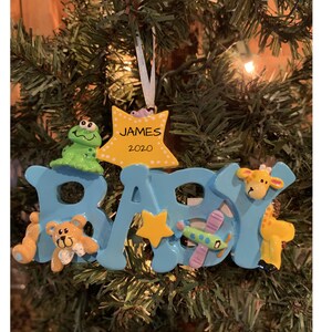May include: Blue "BABY" ornament with a yellow star and various animal figures, including a teddy bear, a frog, a giraffe, and a plane. The ornament also includes a yellow star with the text "JAMES 2020".