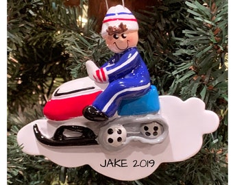 May include: A white, blue, and red snowmobile ornament with a person wearing a blue and white snowsuit and a white hat. The ornament is on a white cloud with the text "JAKE 2019" on the bottom.