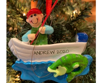 May include: A Christmas tree ornament featuring a fisherman in a white boat with a green fish. The fisherman wears a red hat and green vest. The boat has the name "ANDREW 2020" on the side. The fish is green with yellow accents.