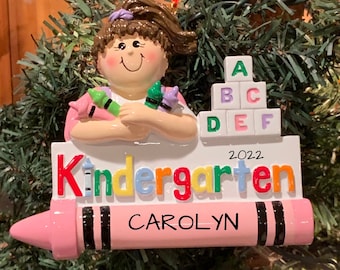 Könnte beinhalten: Rosaer Crayon-Anhänger mit dem Wort "Kindergarten" und dem Jahr "2022" darauf. Der Anhänger zeigt auch ein Mädchen, das Stifte hält und einen Stapel Alphabetblöcke. Der Name "CAROLYN" ist auf den Crayon geschrieben.