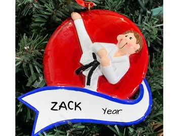 May include: A red and white Christmas ornament featuring a person in a white karate uniform performing a kick. The ornament has a blue and white banner with the name "ZACK" and the word "Year".