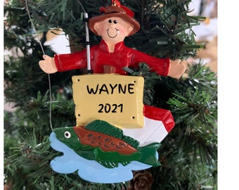 May include: A personalized Christmas ornament featuring a cartoon fisherman holding a fishing rod and a sign that reads "WAYNE 2021". The fisherman is sitting on a blue wave with a green and brown fish.