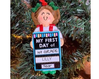Könnte beinhalten: Ein personalisierter Weihnachtsbaumschmuck mit einem Mädchen, das ein Schild mit der Aufschrift "My First Day of 1st Grade Lilly Year" hält. Der Schmuck ist mit bunten Streifen und Sternen verziert.