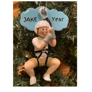 May include: A Christmas tree ornament depicting a person on a zip line. The ornament features a smiling figure wearing a white helmet, gloves, and harness. The text "JAKE Year" is on a blue cloud above the figure.