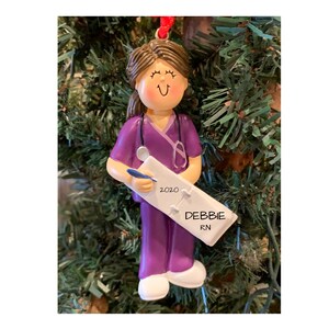 K&ouml;nnte beinhalten: Lila und wei&szlig;er personalisierter Weihnachtsbaumschmuck mit einer Krankenschwester, die eine Klemmbrett mit dem Text "2020" und "DEBBIE RN" h&auml;lt.