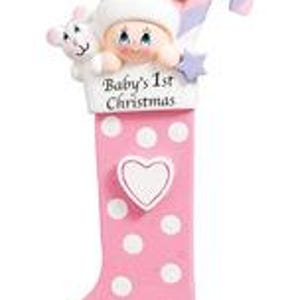 May include: Pink and white polka dot stocking ornament with a baby girl's face peeking out of the top. The stocking has a white heart with a blank space for personalization and the text "Baby's 1st Christmas".