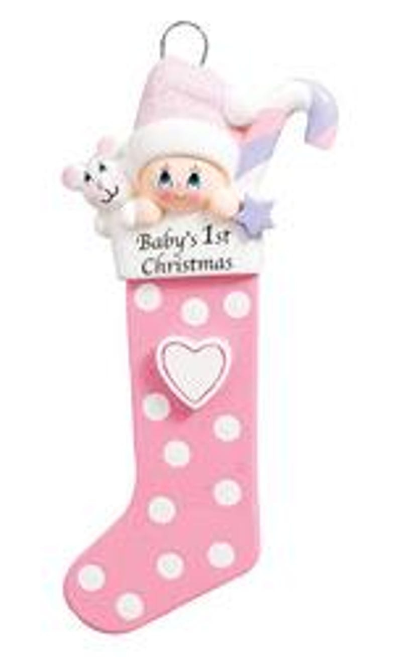 May include: Pink and white polka dot stocking ornament with a baby girl's face peeking out of the top. The stocking has a white heart with a blank space for personalization and the text "Baby's 1st Christmas".