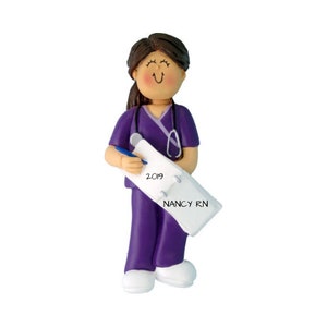 K&ouml;nnte beinhalten: Lilafarbener Scrubs-Anh&auml;nger mit Stethoskop, Stift und Notizblock, auf dem "2019" und "NANCY RN" steht.