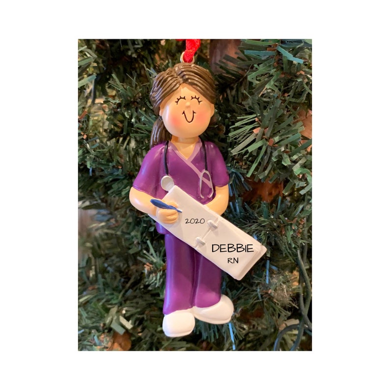 K&ouml;nnte beinhalten: Lila und wei&szlig;er personalisierter Weihnachtsbaumschmuck mit einer Krankenschwester, die eine Klemmbrett mit dem Text "2020" und "DEBBIE RN" h&auml;lt.