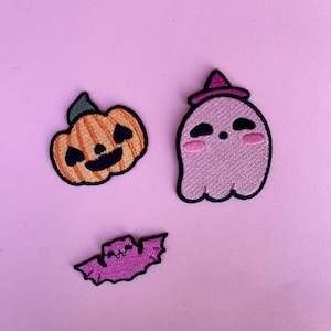 May include: Three embroidered patches featuring a jack-o'-lantern with an orange body and green stem, a light purple ghost wearing a dark purple witch hat with pink cheeks, and a small purple bat with a smiling face and black outlines.