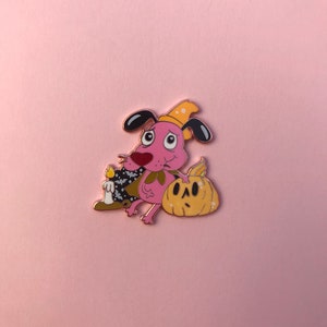 May include: A metal enamel pin featuring a pink dog character wearing a yellow hat and a black cape with white bat patterns. The dog holds a lit candle in one hand and rests the other on a carved yellow pumpkin with a surprised facial expression.